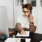 A woman working remotely from home, engaging in a phone call while taking notes.